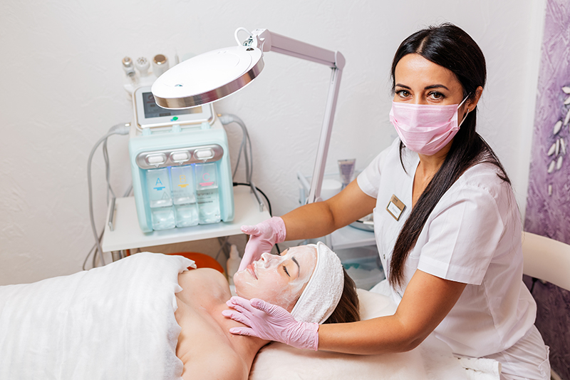 A cosmetologist in a medical mask and rubber gloves applies a cream mask to the client's face. Top view. In the background, a cosmetology device. Concept of cosmetology during the pandemic